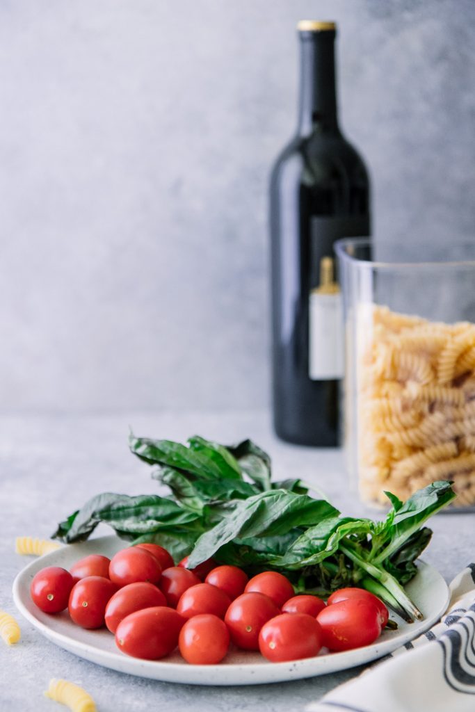 一块白色板材用西红柿和蓬蒿在一张蓝色桌上与一个瓶子fusilli面团和一瓶酒在背景中。