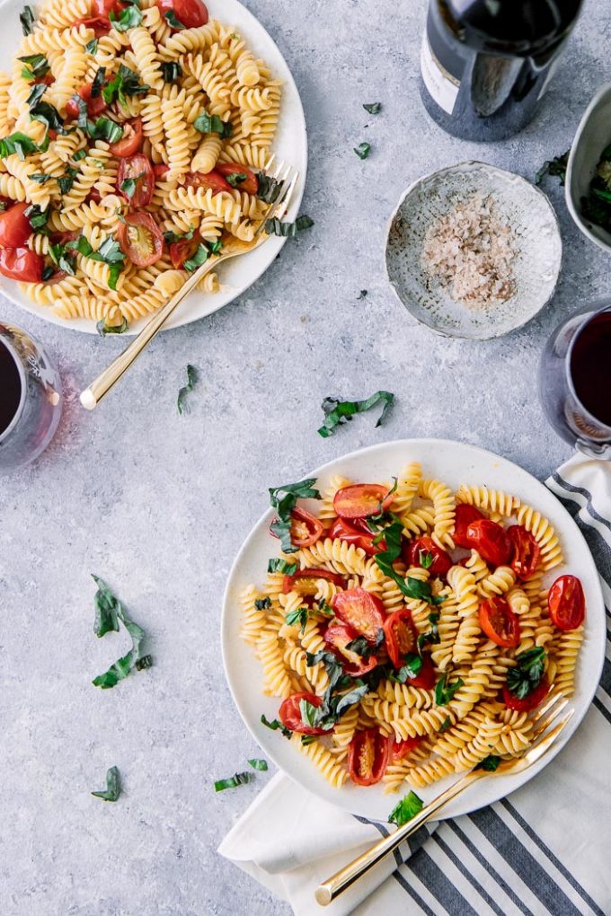 意大利面沙拉两块白色板材用蕃茄，蓬蒿和fusilli面团在一张蓝色桌上与一杯酒。