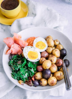 一个早午餐碗，里面有三文鱼、土豆、绿色蔬菜、蒜泥蛋黄酱和鸡蛋，放在白色的桌子上，还有一个黄色的浓缩咖啡杯。