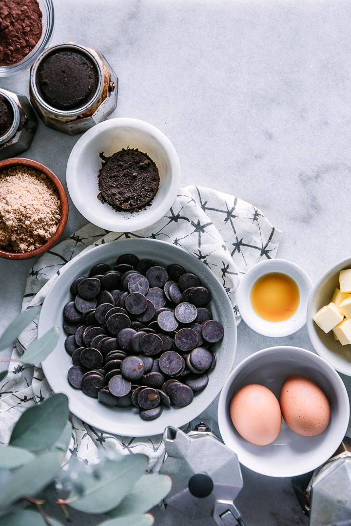 一碗巧克力片，两个鸡蛋，一个香草模子，切好的黄油，红糖和咖啡渣放在一张白色的桌子上，还有一张黑白相间的餐巾