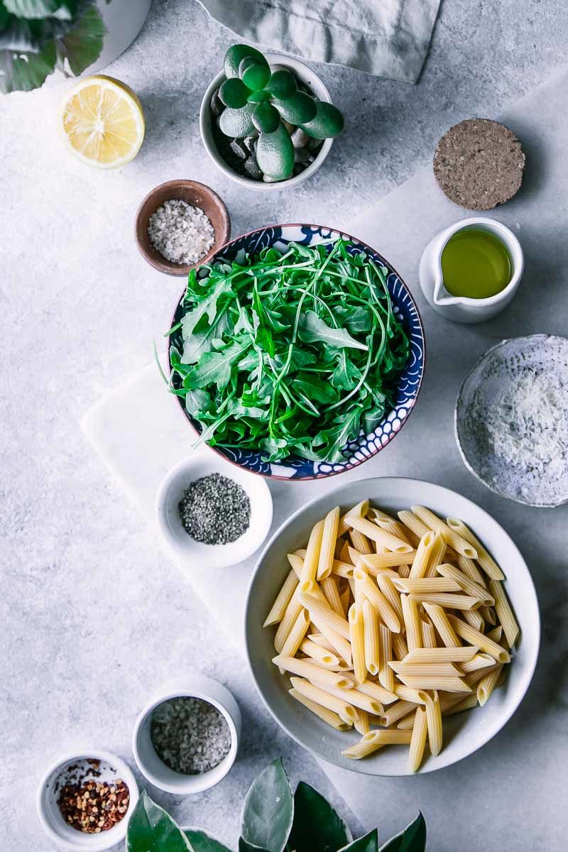 一碗通心粉、芝麻菜、辣椒片、橄榄油、海盐和帕尔马干酪放在白色的桌子上，桌上放着一棵绿色的植物