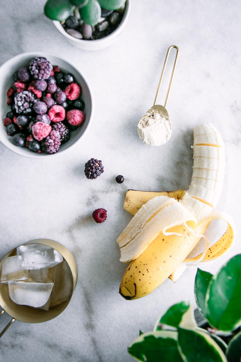 一碗冷冻浆果，一个半被剥皮的香蕉和测量在白色桌上的冰;三重浆果香蕉冰沙的成分