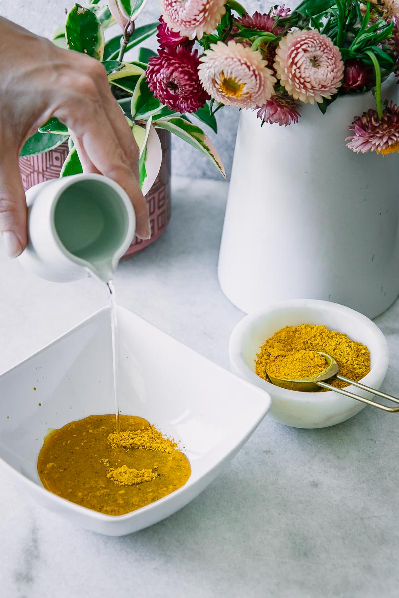 一只手从大罐子里把水倒进一碗白色的碗里，碗里盛着姜黄和金色的牛奶香料