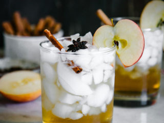 在冰的苹果汁醋闪闪发光洒在鸡尾酒玻璃用苹果切片和肉桂条在与词的一张白色桌上装饰