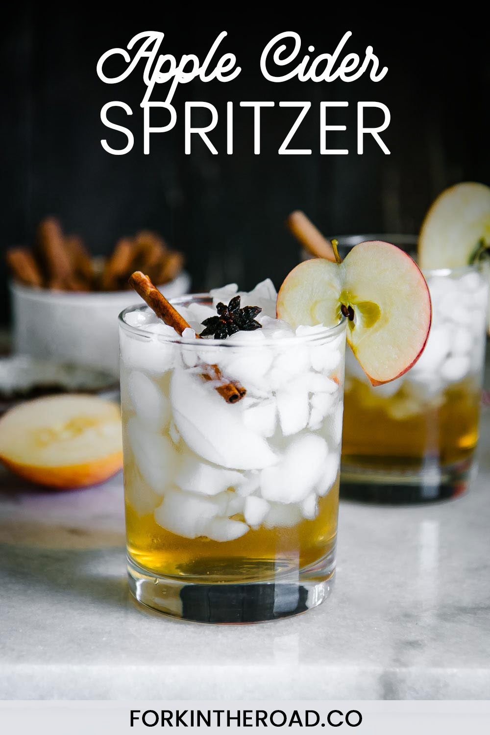 在冰的苹果汁醋闪闪发光洒在鸡尾酒玻璃用苹果切片和肉桂条在与词的一张白色桌上装饰