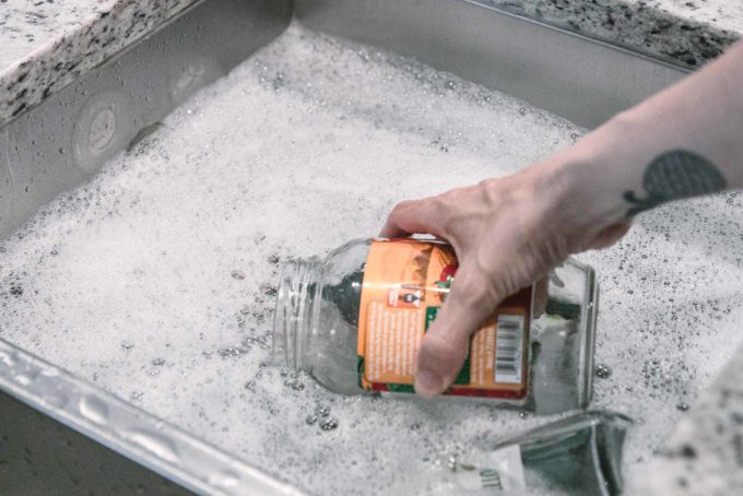 一只手将一个贴着标签的空食品玻璃罐放入装满肥皂水的水槽中
