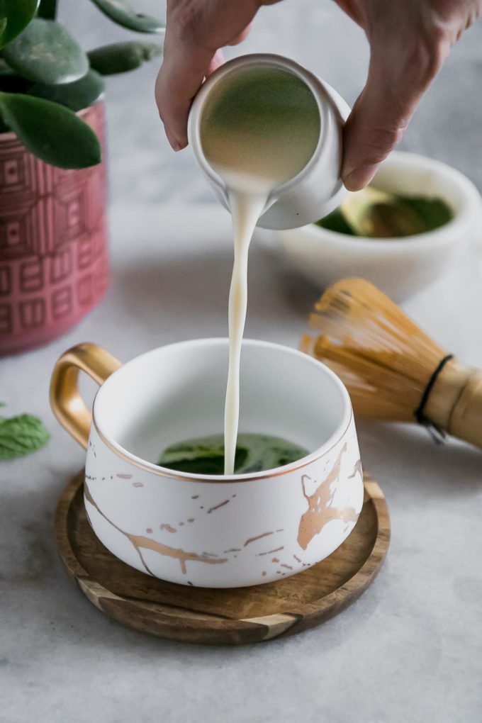把牛奶倒进白色陶瓷杯子里的手，杯子里盛满了抹茶绿茶